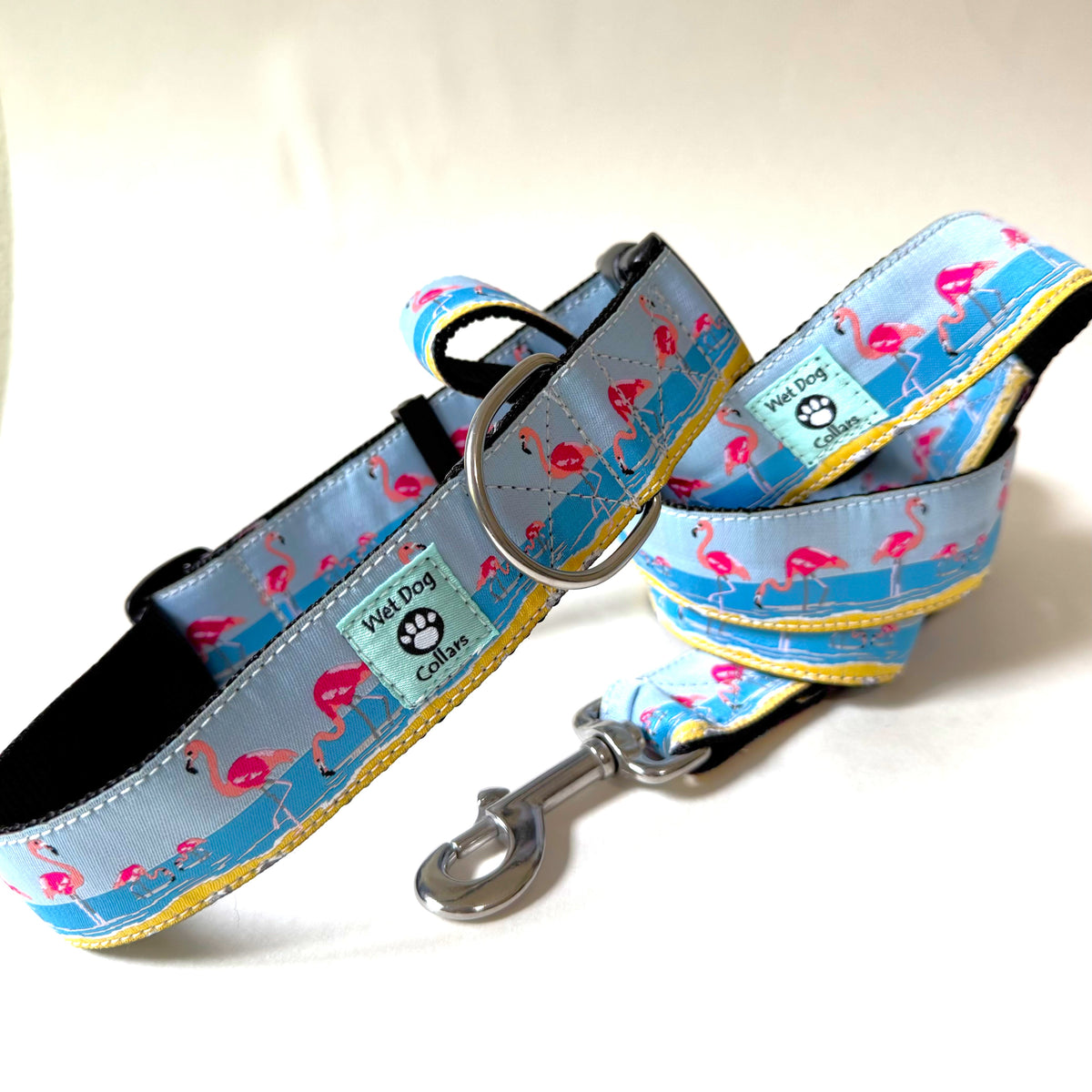 A dog collar and matching leash set with a light blue background, pink flamingo, and sand border, shown on a clean white background. The collar has a stainless metal ring and buckle, and the leash has a stainless metal clasp.