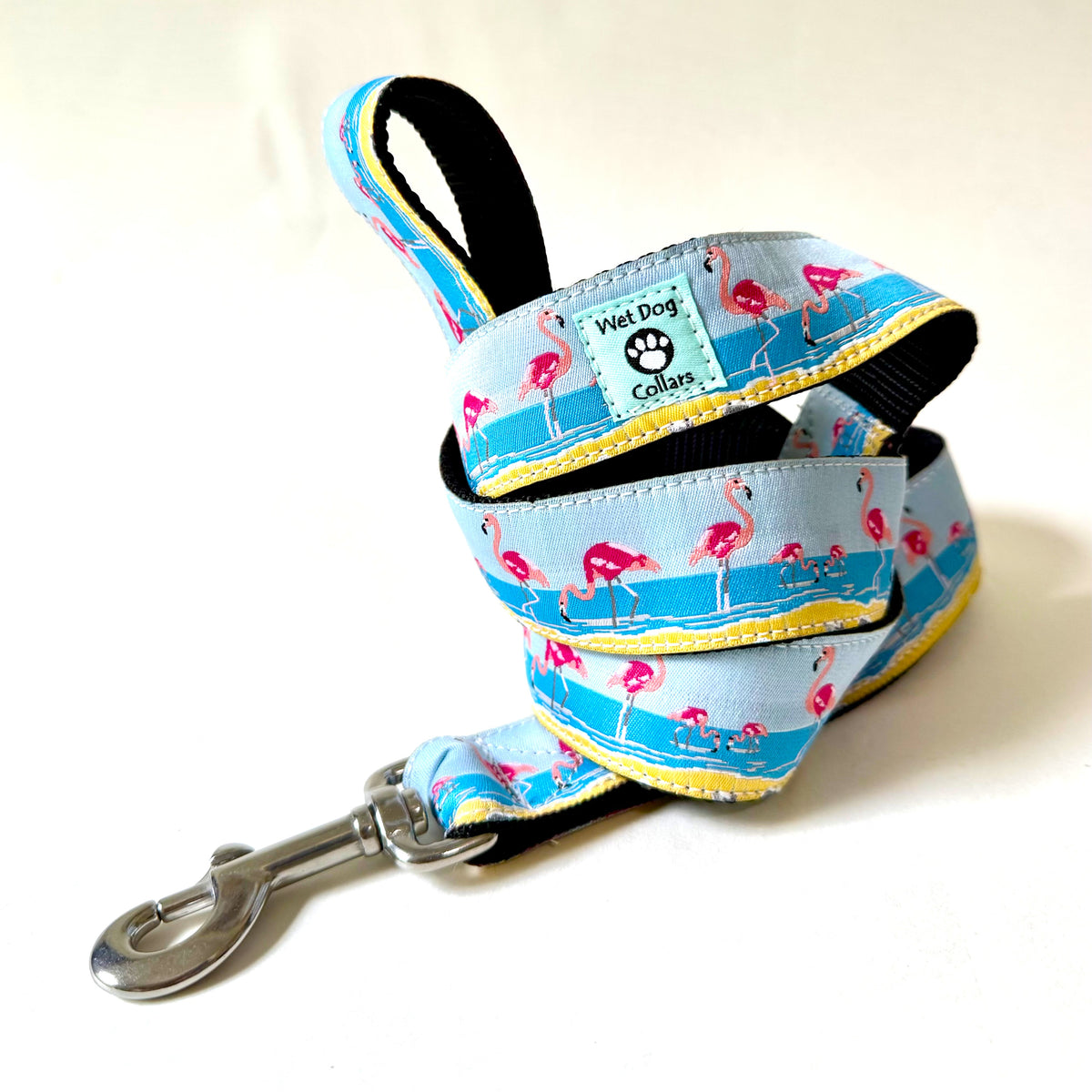 A coiled dog collar with a rust proof metal clasp, featuring a light blue ocean with pink flamingos, with black webbing and a yellow stitched edge. A brand label reads 'Wet Dog Collars'.