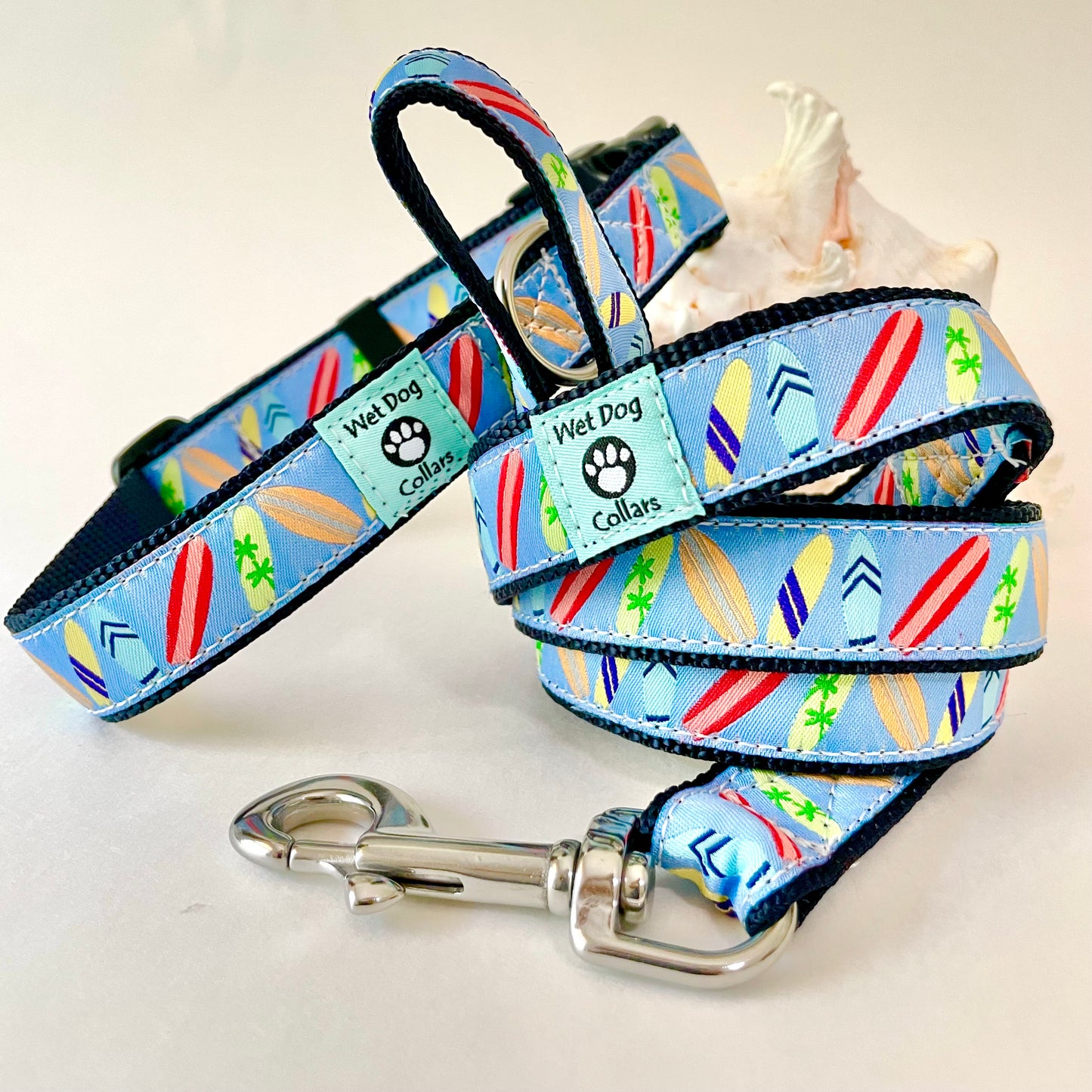 A light blue dog collar and matching leash with a colorful surfboard pattern, displayed on a white background with a small seashell prop. The collar and leash have black trim and a silver metal clasp.