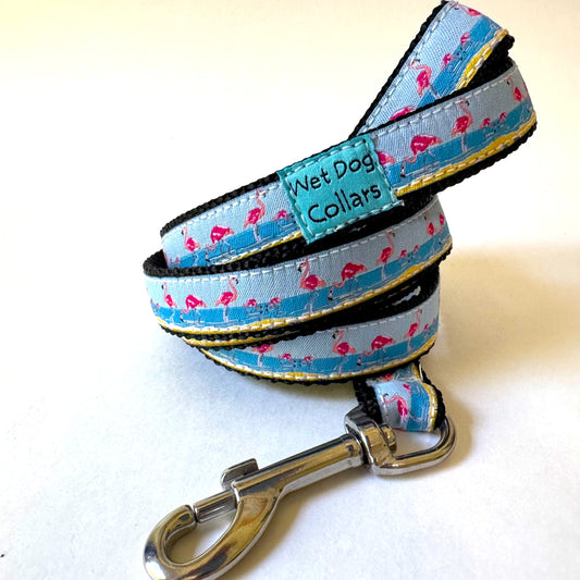 A coiled dog collar with a rust proof metal clasp, featuring a light blue ocean with pink flamingos, with black webbing and a yellow stitched edge. A brand label reads 'Wet Dog Collars'.