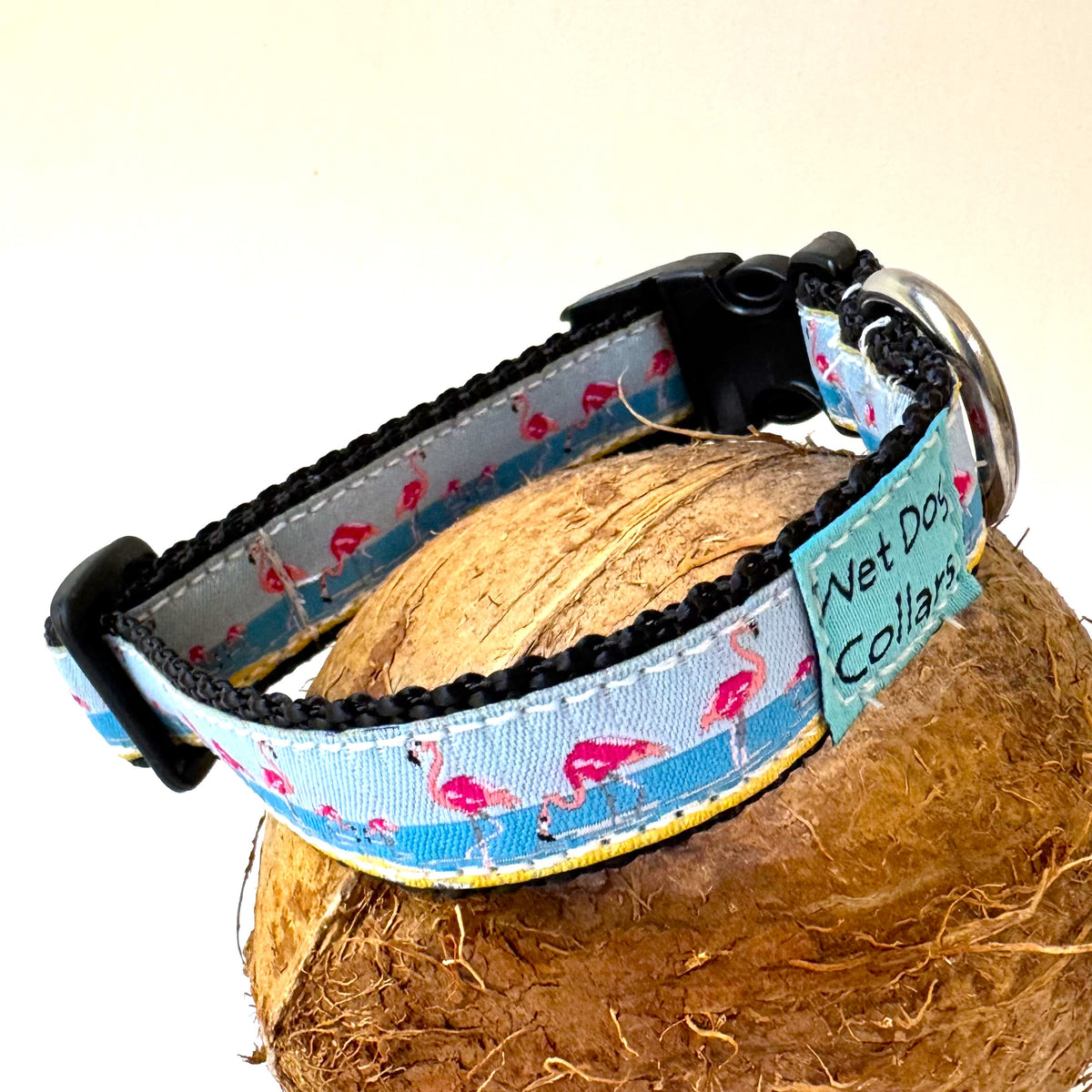 A dog collar with a light blue and pink flamingo pattern, displayed on a brown coconut shell. The collar features black plastic hardware, a stainless D ring, and a blue fabric tag that reads 'Wet Dog Collars'.