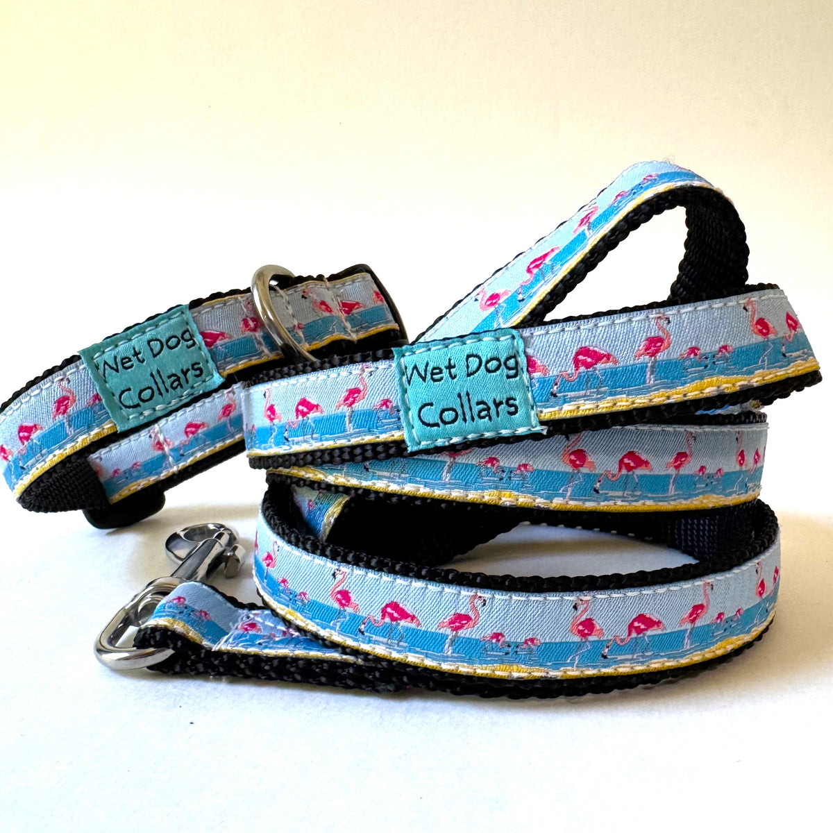 A set of dog collars and leashes from Wet Dog Collars, featuring a light blue and white patterned fabric with pink flamingos and yellow stripes, black webbing, and rust proof metal hardware.