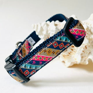 A woven dog collar with a colorful geometric pattern in pink, orange, yellow, and blue on a navy base, displayed on a white seashell against a light background.