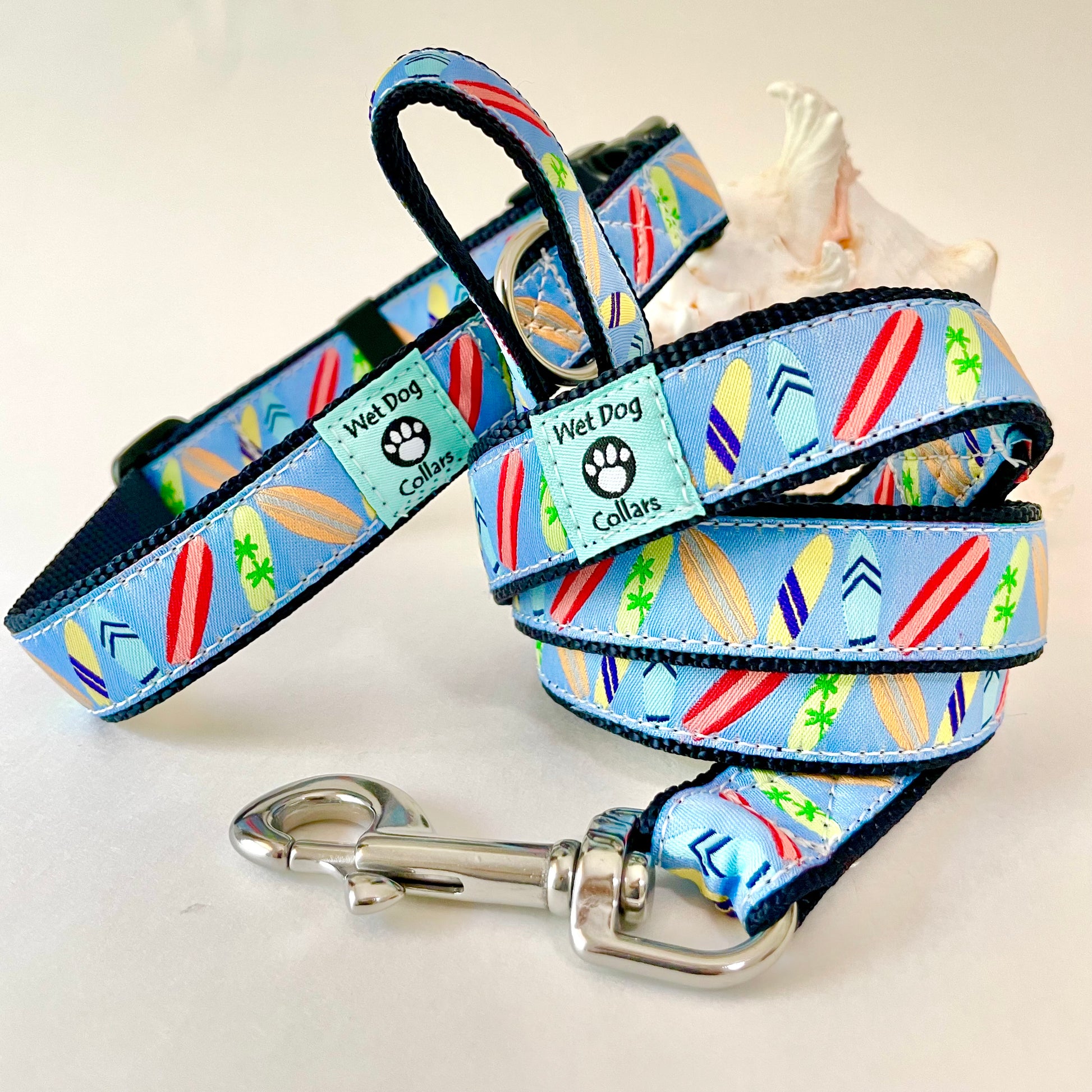 A light blue dog collar and matching leash with a colorful surfboard pattern, displayed on a white background with a small seashell prop. The collar and leash have black trim and a silver metal clasp.