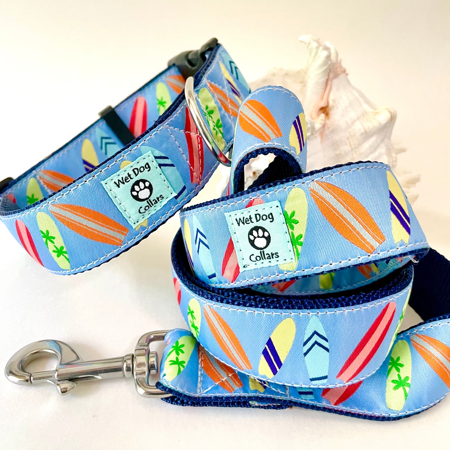 A blue dog collar and matching leash with a colorful surfboard print. The collar and leash have a light blue base with orange, yellow, red, green, and white surfboards, and a light blue brand patch that reads 'Wet Dog Collars' with a paw print.